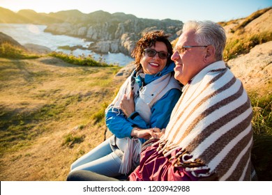 Loving mature couple hiking, sitting on windy top of rock, exploring. Active Mature man and woman wrapped in blanket, hugging and Happily smiling. Scenic view of sea, mountains. Norway, Lindesnes. - Powered by Shutterstock