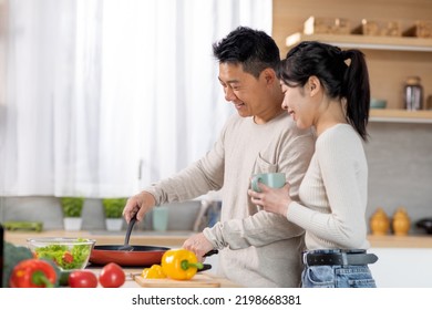 Loving Middle Aged Chinese Man Husband Cooking Delicious Healthy Dinner For His Beautiful Young Wife, Happy Asian Spouses Having Conversation While Preparing Food At Kitchen, Copy Space