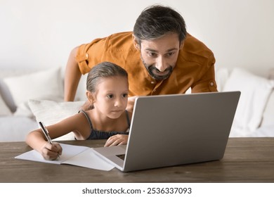 Loving middle aged Hispanic dad helping little daughter kid with elementary school homework, pointing at laptop, standing near child writing notes in copybook, watching video class - Powered by Shutterstock