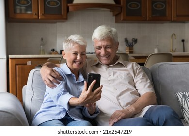 Loving Middle Aged Hoary Married Couple Hug On Sofa At Own Studio Apartment Shoot Family Selfie On Cellphone Make Videocall Use Funny App. Smiling Senior Spouses Enjoy Watching Video At Social Network