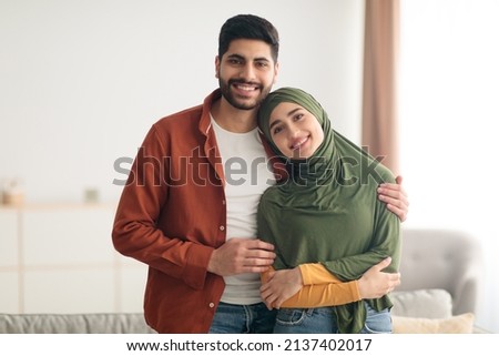 Loving Middle Eastern Couple Embracing Posing Standing At Home. Muslim Husband Hugging His Wife With Hijab Smiling To Camera. Love And Romance, Happy Relationship And Marriage Concept