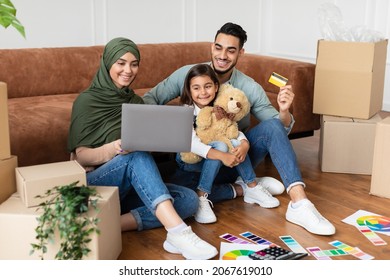 Loving Middle Eastern Family Using Laptop And Debit Credit Card, Choosing Furniture After Moving To New Home, Happy Parents And Kid With Computer Purchasing In Internet, Sitting On Floor