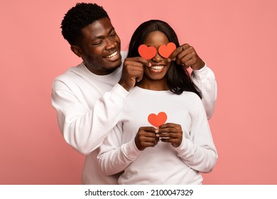 Loving Millennial Black Guy Covering His Happy Girlfriend Eyes With Red Valentine Cards And Smiling, Happy African American Couple Celebrating Valentine Day Together On Pink Studio Background