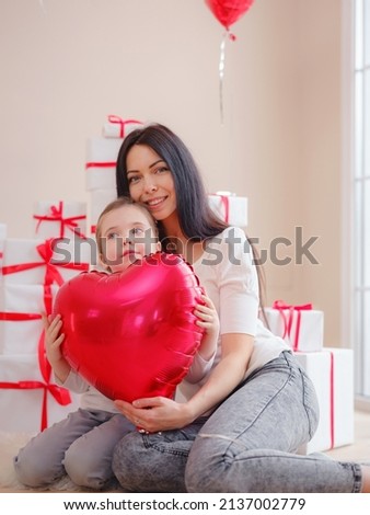 Loving mom hug little daughter hold red heart shaped balloons , express love and gratitude with pleasant words and best wishes at birthday, family smiling. Mother or Valentine's day celebration