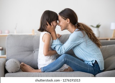 Loving mom laughing caressing touching noses with cute kid daughter sitting on sofa at home, happy mother and little child girl hugging having fun playing enjoy funny moments good time together