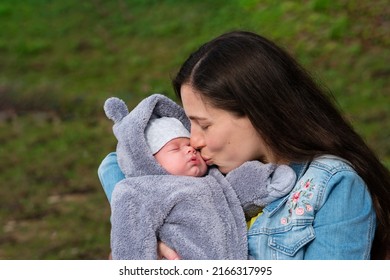 Loving Mom with Newborn Son on Her Arms in Spring Park. Сareful Mommy Kissing at Cute Sleeping New Born Child. Family Spend Time Together on Nature Outdoors. First Month Baby Life