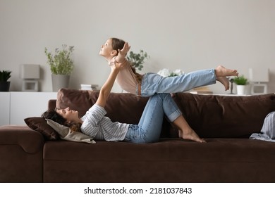 Loving mom play with little kid at home, lying on sofa lifts on arms her cute 6s daughter feeling freedom imagining like flying in air. Active playtime leisure, daydreams about family vacation concept