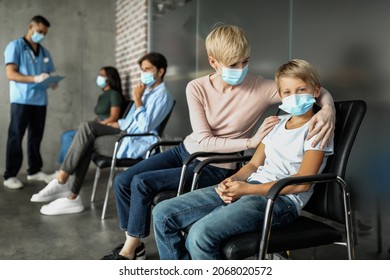 Loving Mother Blonde Woman Comforting Her Teen Son While Waiting For Vaccination Against Coronavirus At Clinic, Multiracial Group Of Patients Sitting In Queue, Having Conversation With Doctor