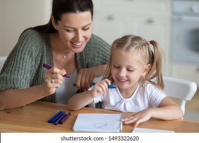 Loving mother and daughter sitting at table having good free time drawing together with colorful felt-tip pens on exercise book, parent helping to child with picture hobby and leisure activity concept