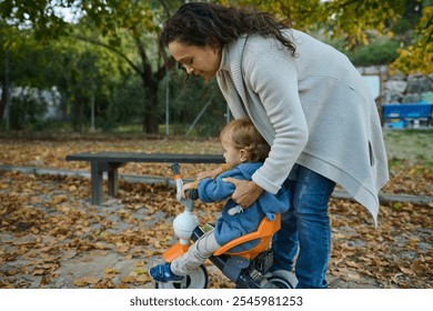 A loving mother helps her young child ride a tricycle in a park filled with autumn leaves, symbolizing care and family bonding in a serene outdoor environment. - Powered by Shutterstock