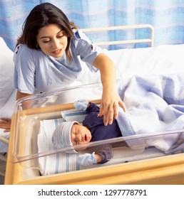 Loving Mother Looking At Down At Newborn Baby Boy In Hospital Crib