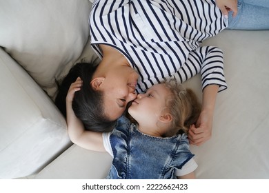 Loving Mother Lying On Couch Touching Nose Face With Little Daughter At Home. Top View Mom And Kid Girl Relaxing On Sofa, Enjoy Cute Moment Together. Happy Motherhood, Family Bond, Unconditional Love.