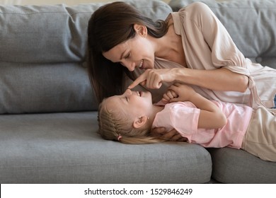 Loving mother playing with little daughter, touching nose, lying on couch at home, happy young mum and adorable preschool child girl having fun together on weekend, enjoying tender moment