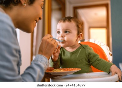 Loving mother spoon-feeding her happy baby boy. Adorable toddler in green outfit enjoying a nutritious meal at home. - Powered by Shutterstock