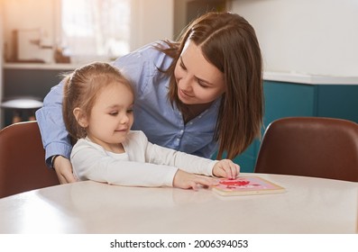 Loving mother and sweet little daughter sitting at table and playing with puzzles together at home