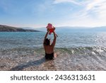Loving mother swimming with toddler on paradise pebble beach in coastal town Baska, Krk Island, Primorje-Gorski Kotar, Croatia, Europe. Scenic view of coastline of Mediterranean Adriatic Sea in summer