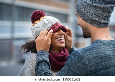 Loving Multiethnic Couple Having Fun Outdoor In Winter. Young Man Covering Eyes His Girl With Woolen Cap. Cheerful Guy And Smiling African Woman Playing In Winter Outdoor.