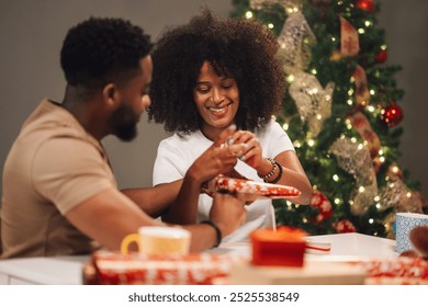 A loving multiracial couple wraps Christmas gifts together, exchanging smiles. The room is cozy with decorative elements including a brightly lit tree, capturing festive warmth and cheer. - Powered by Shutterstock