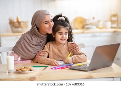 Loving Muslim Mom Helping Her Little Daughter To Study With Laptop At Home, Cute Child And Islamic Mother Sitting At Kitchen Together And Looking At Computer Screen, Enjoying Online Education