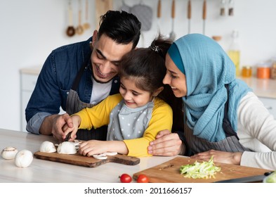 Loving Muslim Parents Teaching Cute Little Girl How To Cook, Small Female Child Chopping Vegetables, Happy Young Middle Eastern Family Of Three Enjoying Cooking Together At Home, Closeup Shot