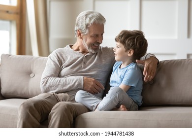 Loving old 60s Caucasian granddad sit on sofa with little curious grandson talk and chat at home. Caring mature grey-haired grandfather relax on couch with small grandchild gossip share secrets.