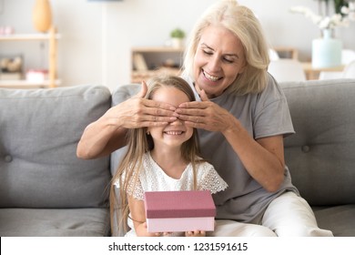 Loving Old Grandmother Making Present To Little Happy Kid Girl Holding Gift Box, Middle-aged Granny Closing Eyes Of Cute Smiling Grandchild Excited With Birthday Surprise From Grandma Sitting On Sofa