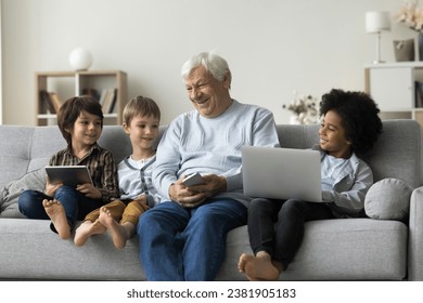 Loving older great-grandpa and great-grandsons sit on couch holding diverse modern devices, laugh, enjoy pastime on internet and communication. Modern technologies overuse, bad habit, leisure online - Powered by Shutterstock