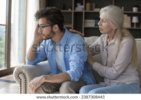 Loving older woman comforting her upset young adult son, gives wise advice in difficult life situation, talking, provide psychological support showing care seated on couch, spend time together at home