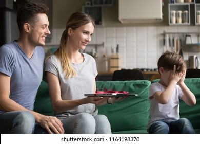 Loving Parents Mom And Dad Making Present To Kid Son Holding New Laptop Presenting As Gift, Cute Child Boy Closing Eyes With Hands Waiting For Birthday Surprise From Mother Father Sitting On Sofa