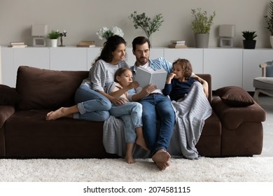 Loving parents with two little kids reading book together, hugging, sitting on cozy couch at home, smiling mother and father in glasses with adorable daughter and son engaged in educational activity - Shutterstock ID 2074581115