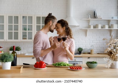 Loving passionate happy young Caucasian family couple touching foreheads, clinking glasses of red wine, enjoying tender affectionate moment together in modern kitchen, distracted from preparing food.