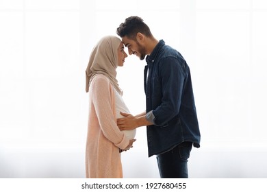 Loving pregnant muslim couple bonding together at home, standing near window, expecting arab family touching foreheads and smiling, romantic husband embracing belly of his wife in hijab, side view
