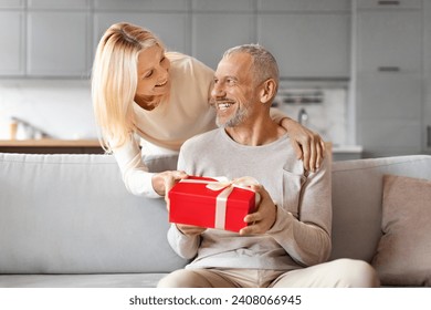 Loving pretty blonde mature woman giving present her surprised husband. Happy senior married couple exchanging gifts on marriage anniversary, celebrating birthday, home interior - Powered by Shutterstock