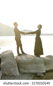 Loving  Relationship. Elegantly Dressed Loving Couple Stand By The Sea At Sunset Holding Their Hands And Looking At Each Other's Eyes. Historical Romance Novel. 19th Century Style.