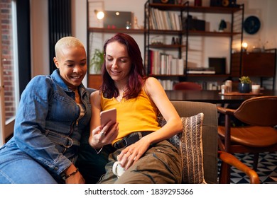 Loving Same Sex Female Couple Sitting On Sofa At Home Looking At Mobile Phone Together