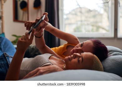 Loving Same Sex Female Couple Lying On Bed At Home Looking At Mobile Phone Together