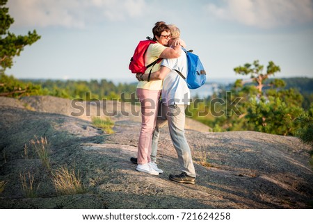 Loving senior couple hiking, standing on the top of rock, exploring. Active Mature man and woman hugging and kissing smiling. Scenic view of gulf and sea on sunset. Healthy lifestyle. Finland.