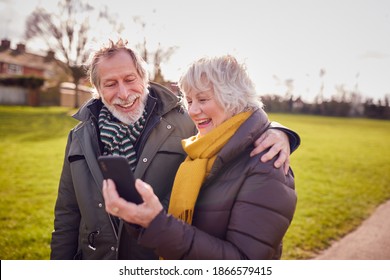 Loving Senior Couple Looking At Mobile Phone Enjoying Autumn Or Winter Walk Through Park Together