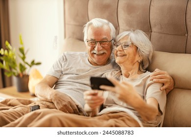 Loving senior couple is sitting on a bed in a cozy bedroom at home and using cellphone for selfies. - Powered by Shutterstock