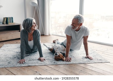 Loving Senior Couple In Sports Clothing Doing Yoga And Smiling While Spending Time At Home With Their Dog