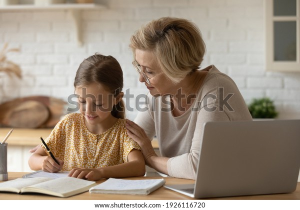 Loving senior grandmother hug shoulders of small\
girl grandchild assist in preparing homework support give advice.\
Friendly aged woman teacher watching little kid pupil writing maths\
problem solution