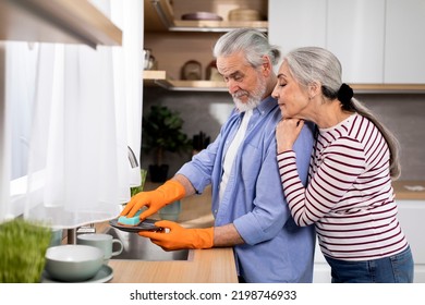 Loving Senior Woman Embracing Her Husband From The Back While He Washing Dishes In Kitchen, Happy Elderly Spouses Sharing Domestic Chores, Caring Husband Helping With Cleaning, Side View