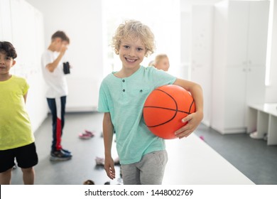 Loving Sports. Smiling Pupil Holding His Big Basketball Standing In Changing Room With His Classmates.