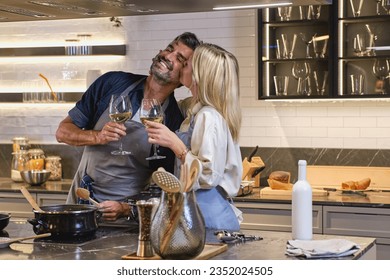 Loving wife kissing husband on cheek while cooking together at home and standing with glasses of white wine at stove - Powered by Shutterstock