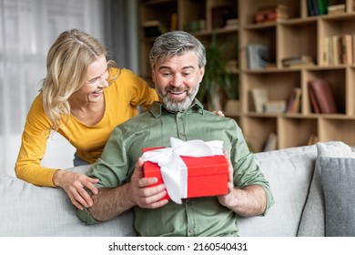 Loving wife surprising her middle aged husband with gift at home, greeting him with birthday or anniversary, happy woman giving present box to excited man, celebrating valentine's day together - Powered by Shutterstock