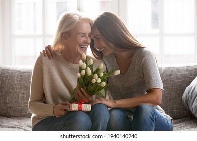 Loving young adult female child congratulate excited elderly mother with birthday anniversary at home. Smiling caring grownup millennial daughter present gift flowers to old mom on women s day.