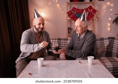 Loving young adult son congratulate elderly father with anniversary lighting candles on cake at home. Smiling millennial man present cake to old dad on his birthday. Copy space - Powered by Shutterstock