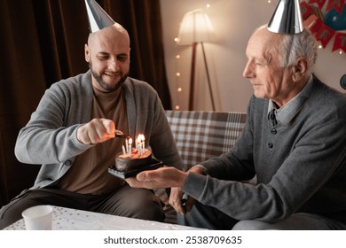 Loving young adult son congratulate elderly father with anniversary lighting candles on cake at home. Smiling millennial man present cake to old dad on his birthday. Copy space - Powered by Shutterstock