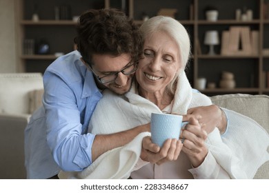 Loving young adult son giving positive sick senior mother help, assist, care, wrapping mom shoulders into plaid, scarf, giving cup of hot drink, hugging, smiling, laughing - Powered by Shutterstock