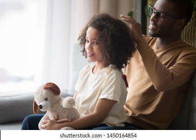 Loving young african American dad sit on couch with preschooler daughter brush curly hair, caring black father spend time with girl child take care making hairdo relaxing at home together - Powered by Shutterstock